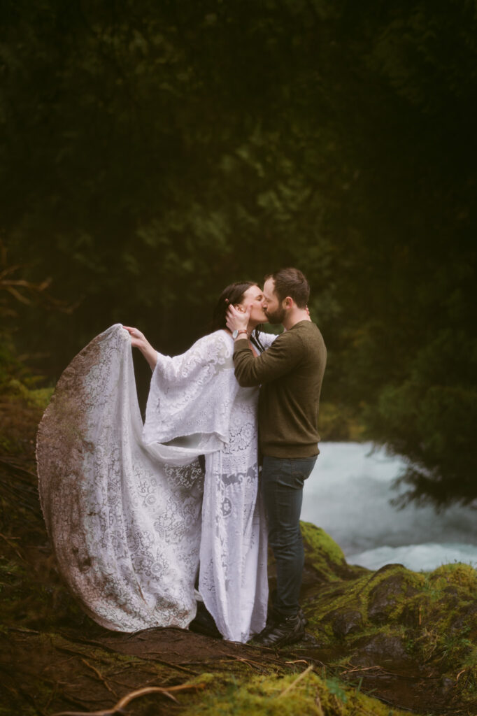 A bride and groom stand at the base of Sahalie Falls in Oregon. The groom holds the bride's face, kissing her, as the bride swings the train of her dress back and forth.