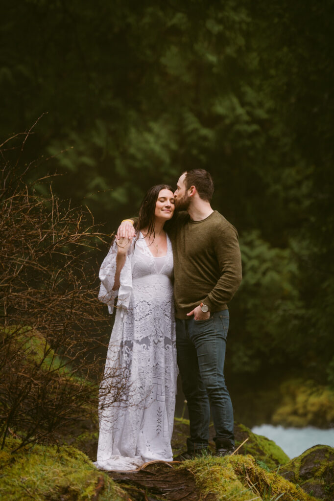 A bride and groom stand at the base of Sahalie Falls in Oregon. The groom has one hand over the bride's shoulder, the other in his pocket. The bride is holding the groom's hand and leaning in as he kisses her on the temple.