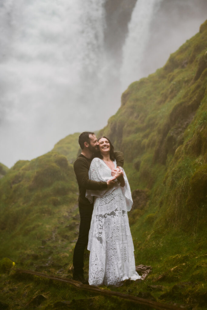 A bride and groom stand at the base of Sahalie Falls in Oregon. The groom is standing behind the bride with his arms wrapped around her while the bride holds his hands. Both are laughing as they get soaked with the spray coming off of the falls.