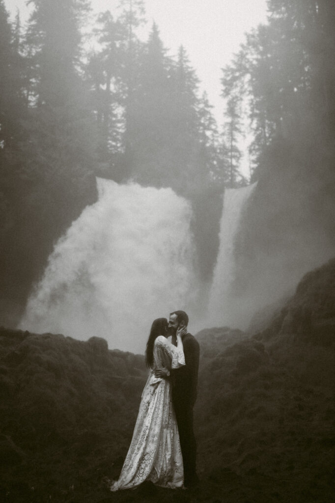 A black and white image of a bride and groom standing at the base of Sahalie Falls in Oregon. The groom is holding the bride's waist as her hands graze his jawline, kissing.