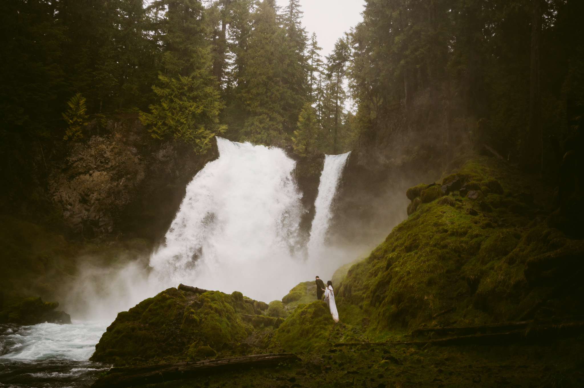 A pulled back landscape shot showing Sahalie falls with a small bride and groom walking towards it.