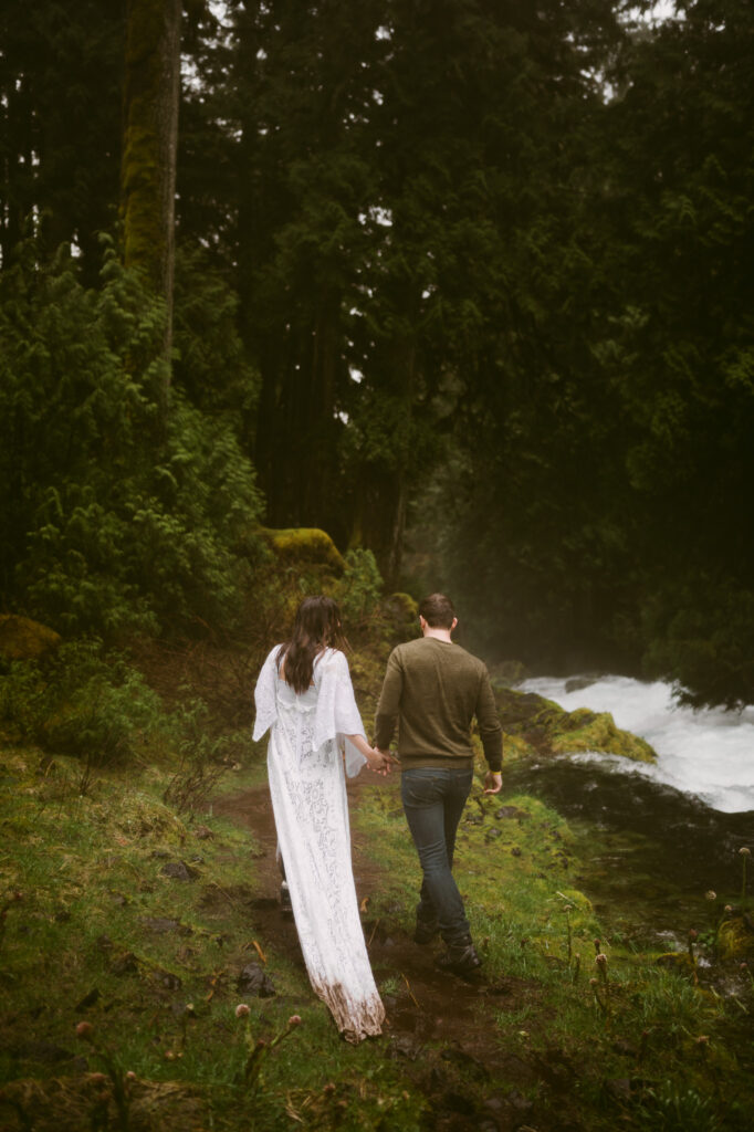 A bride and groom walk hand in hand away from the camera, along a raging pacific northwest stream.