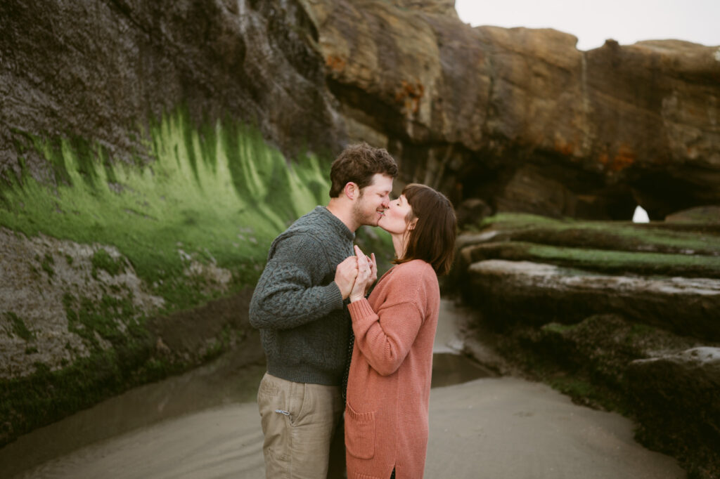 A man and woman holds hands and kiss along the seaweed-covered shoreline of Otter Crest Beach.