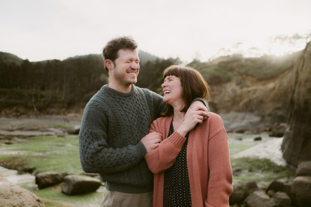 A man and woman laughing together. The woman is holding the man's hand, which is draped over her shoulder.