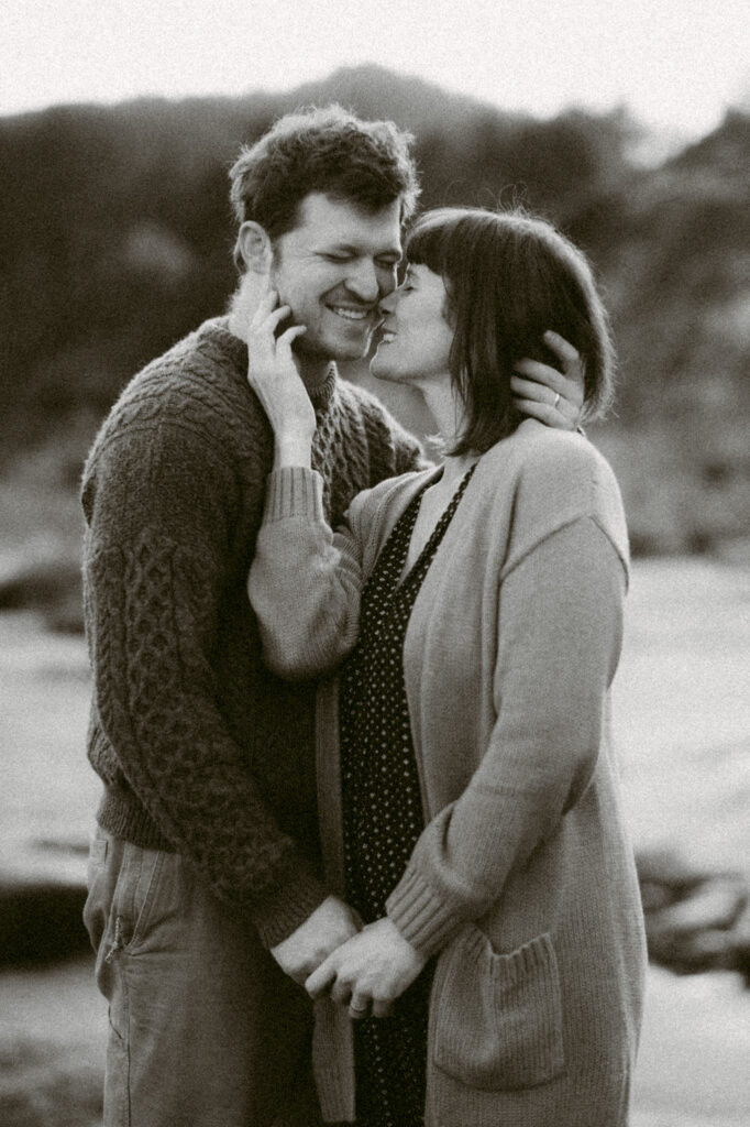 A black and white image of a man and woman at Devil's Punchbowl in Oregon. They are holding hands in front of their bodies. The woman has her other hand on the man's cheek and is kissing his face.