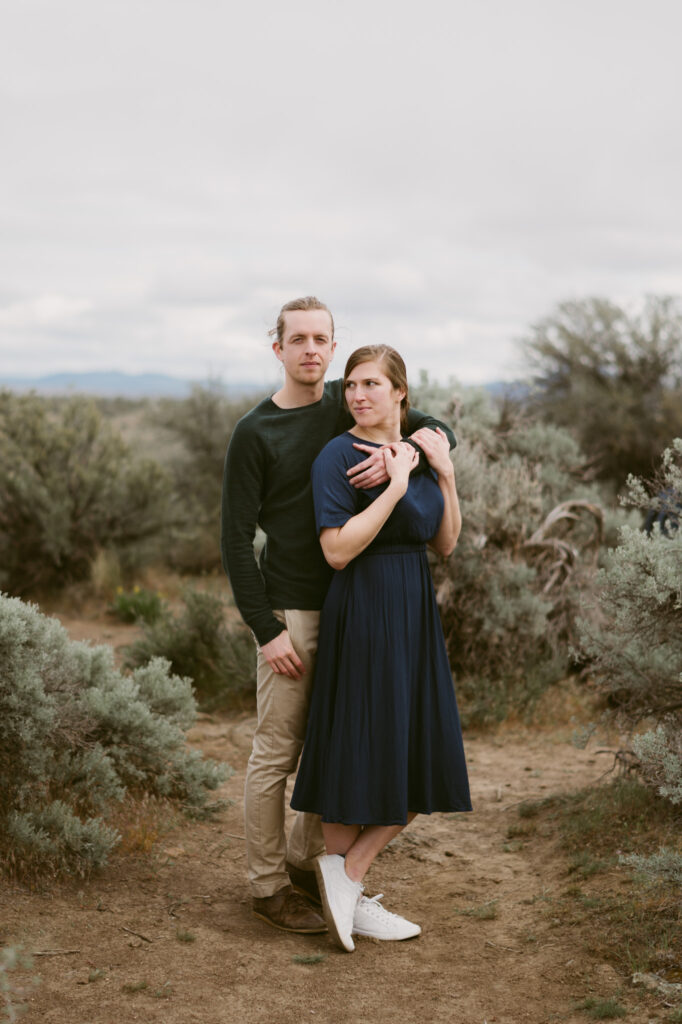 A man and woman stand amongst the sage bushes at Cove Palisades state park. The man has one arm around the woman's chest, the other in his pocket. The woman is holding onto the man's forearm with both hands and looking away from the camera.