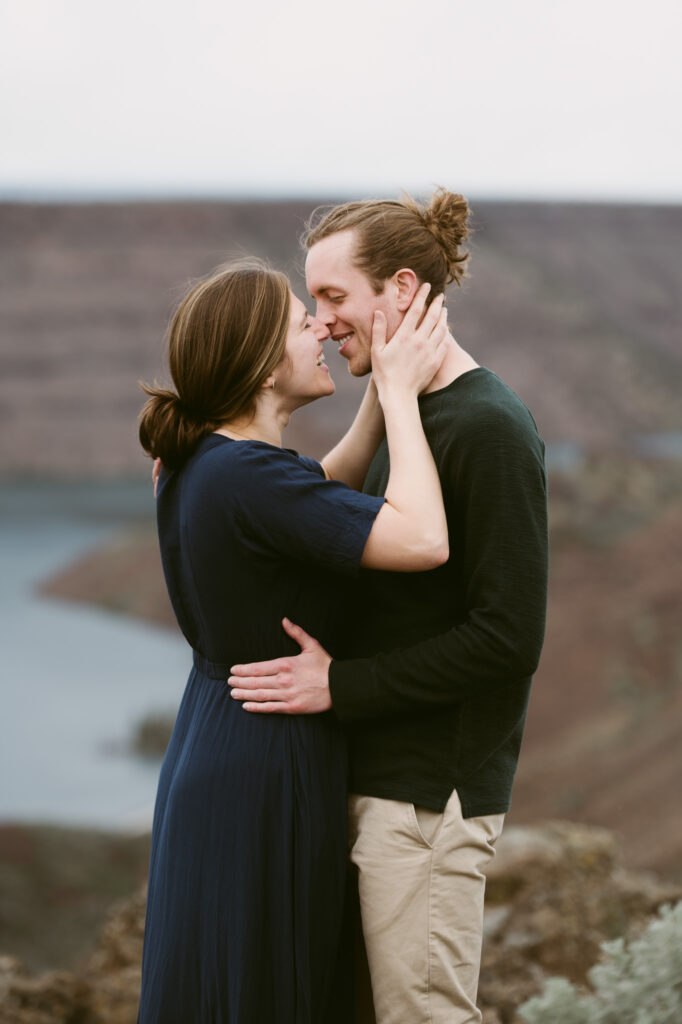 A man and woman stand close together. The man's hands are on the woman's waist while she has her hands on his face, pulling him in for a kiss.