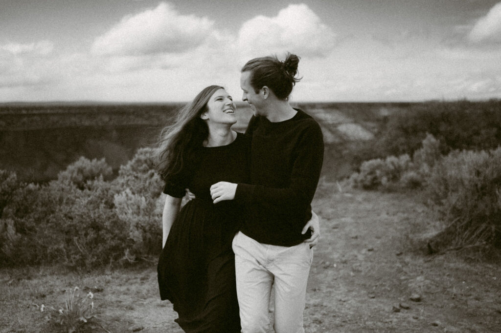 A black and white image of a man and woman walking towards the camera, arms around each other's waists and laughing.