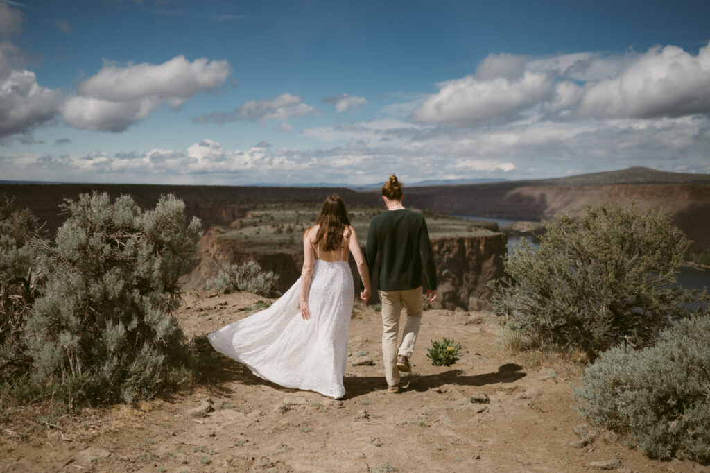 A man and woman walk away from the camera, hand in hand. Around them is the Cove Palisades State Park.