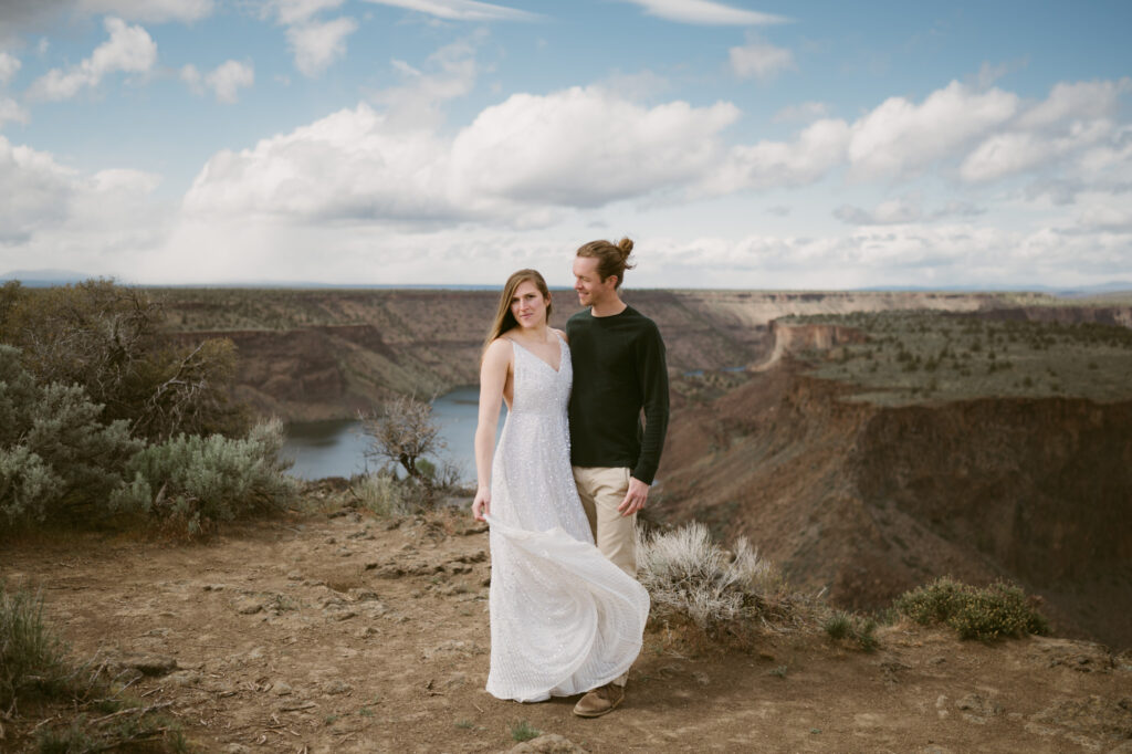 A man and woman stand at the Tam-a-lau Trail overlook at Cove Palisades State Park in Oregon. The woman is wearing a white dress and holding it in front of her so it catches the breeze.