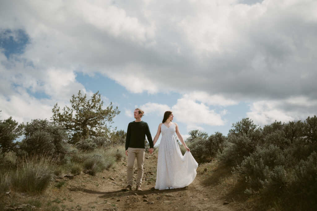 A man and woman stand at the Tam-a-lau Trail overlook at Cove Palisades State Park in Oregon. The woman is wearing a white dress and holding it in front of her so it catches the breeze.