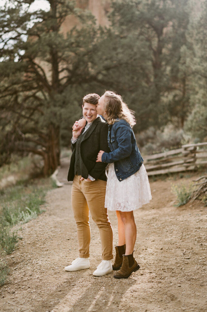 Two women stand with their backs to the sun at Smith Rock State Park in Terrebonne, Oregon. The first woman has her back against the second woman's chest and is holding her hand over her shoulder. The first woman is laughing as the second woman kisses her cheek.