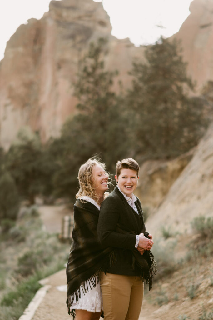 One woman embraces a second woman from behind. The second woman is holding the first woman's hands. Both are laughing.