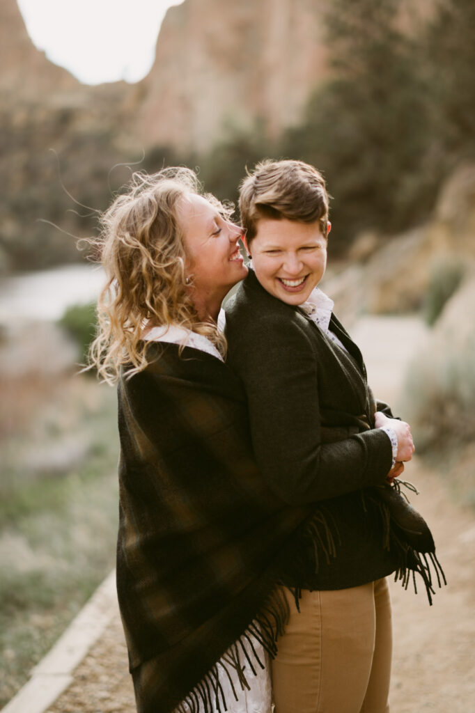 One woman embraces a second woman from behind. The second woman is holding the first woman's hands. Both are laughing.