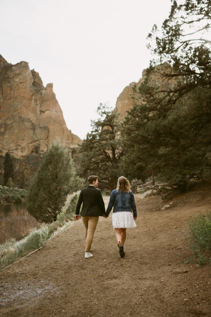 Two women hold hands as they walk away from the camera in Smith Rock State Park, Oregon. One woman is wearing a white sun dress with denim jacket while the other woman is wearing khaki pants, a button down shirt, tweed vest, and jacket.