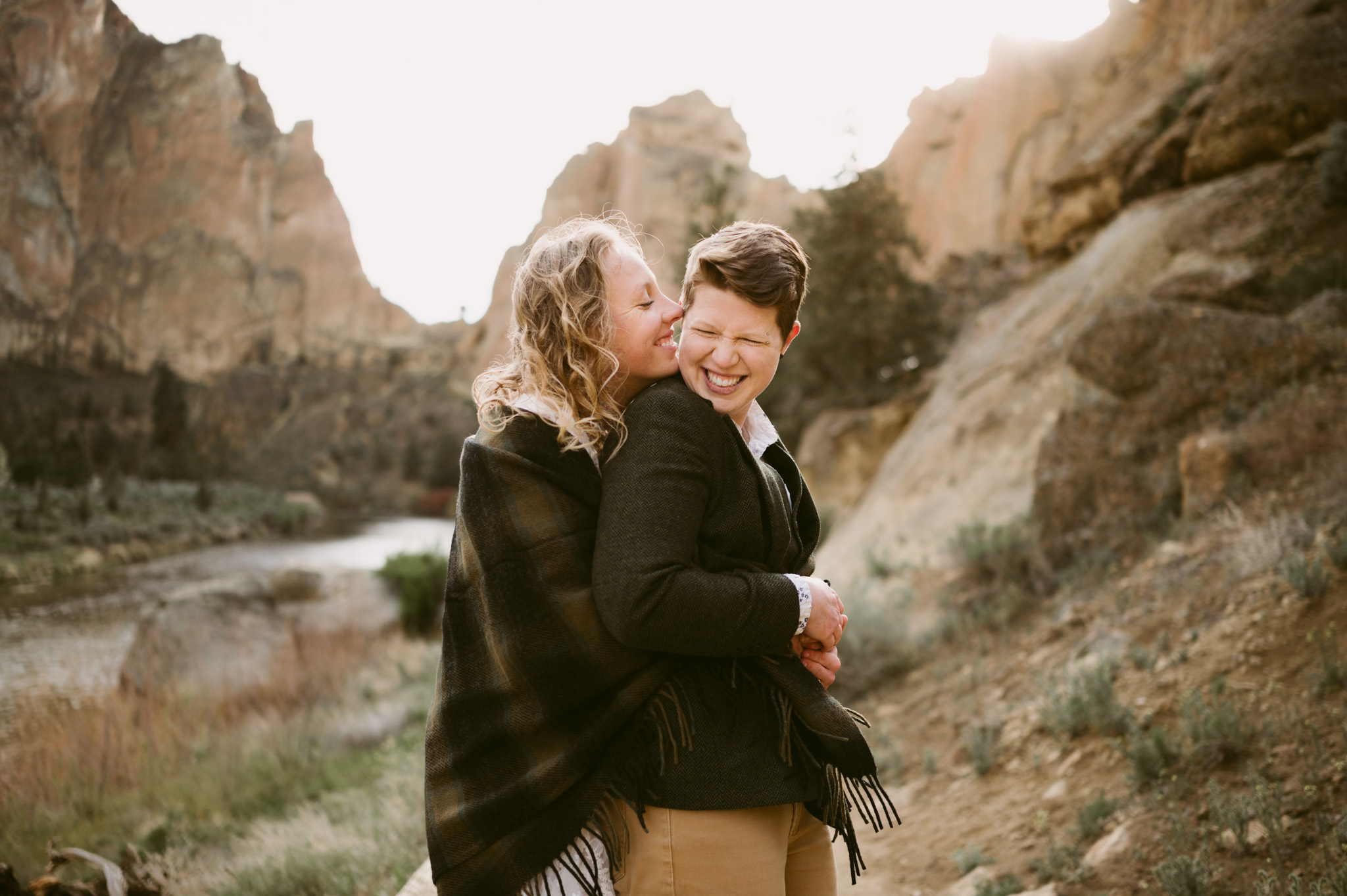 One woman embraces a second woman from behind. The second woman is holding the first woman's hands. Both are laughing.