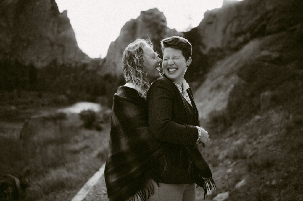 A black and white image where one woman embraces a second woman from behind. The second woman is holding the first woman's hands. Both are laughing.