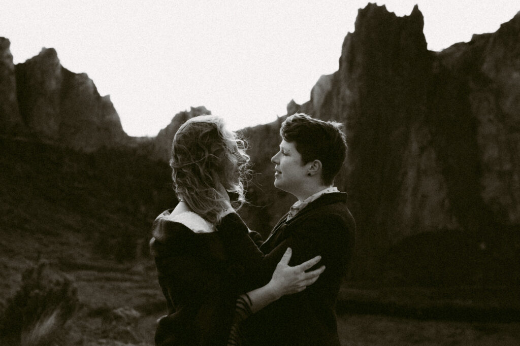 A black and white image of two women staring lovingly into each other's eyes. One woman is holding the arms of the second woman while her face is being caressed by the first woman.