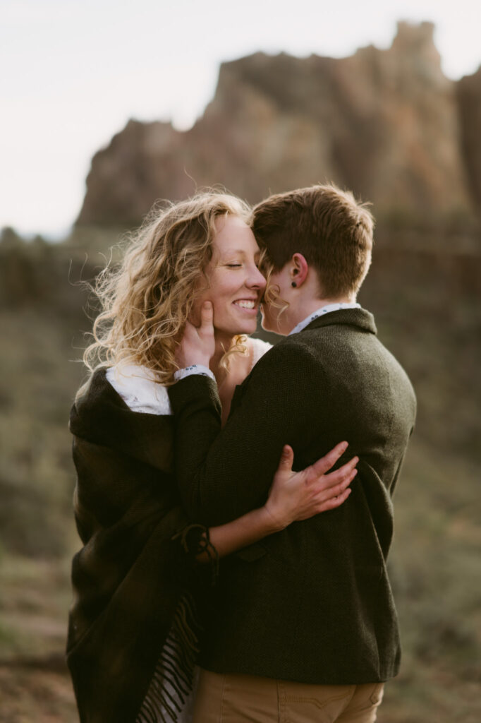 At Smith Rock State Park, two women stand forehead-to-forehead, smiling slightly. The first woman is holding the face of the second woman.