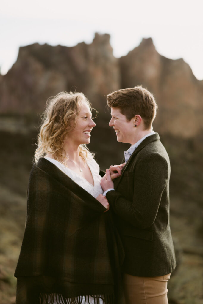 Two women stand facing one another at Smith Rock State Park. They're holding hands between their bodies and are smiling at one another.