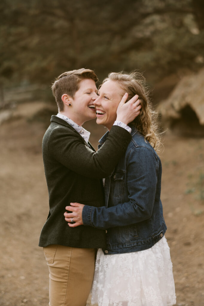 Two women lean in for a kiss. The first woman has her hand on the other's waist. The second woman is tucking the first woman's hair behind her ear as she kisses her cheek.