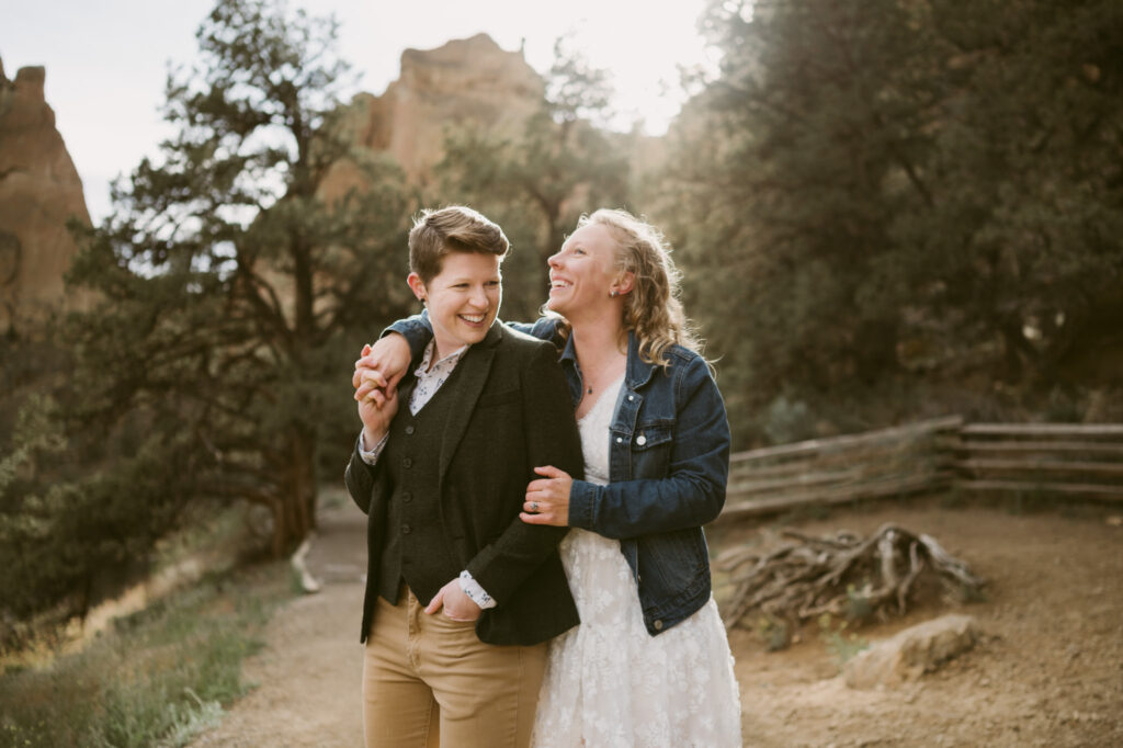 Two women stand with their backs to the sun at Smith Rock State Park in Terrebonne, Oregon. The first woman has her back against the second woman's chest and is holding her hand over her shoulder. They are laughing.