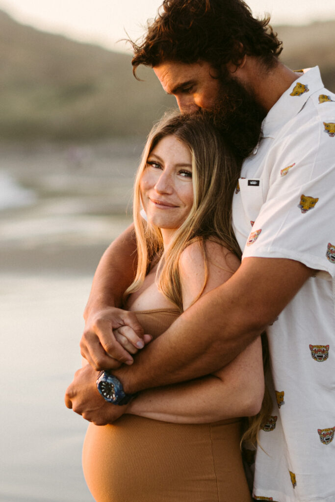 A pregnant woman smiles at the camera as her husband kisses the top of her head at their maternity session at Seal Rock Recreation Site in Seal Rock, Oregon.