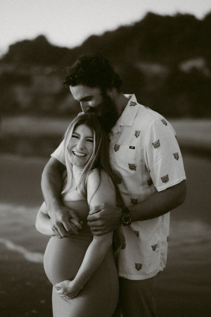 A black and white image of a pregnant woman smiling at the camera as her husband kisses the top of her head at their maternity session at Seal Rock Recreation Site in Seal Rock, Oregon.