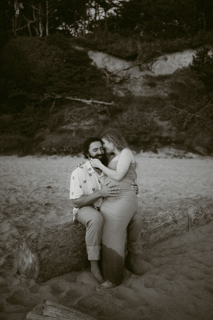 A black and white image of a young couple embracing on a log during their maternity session at Seal Rock Beach in Seal Rock, Oregon.