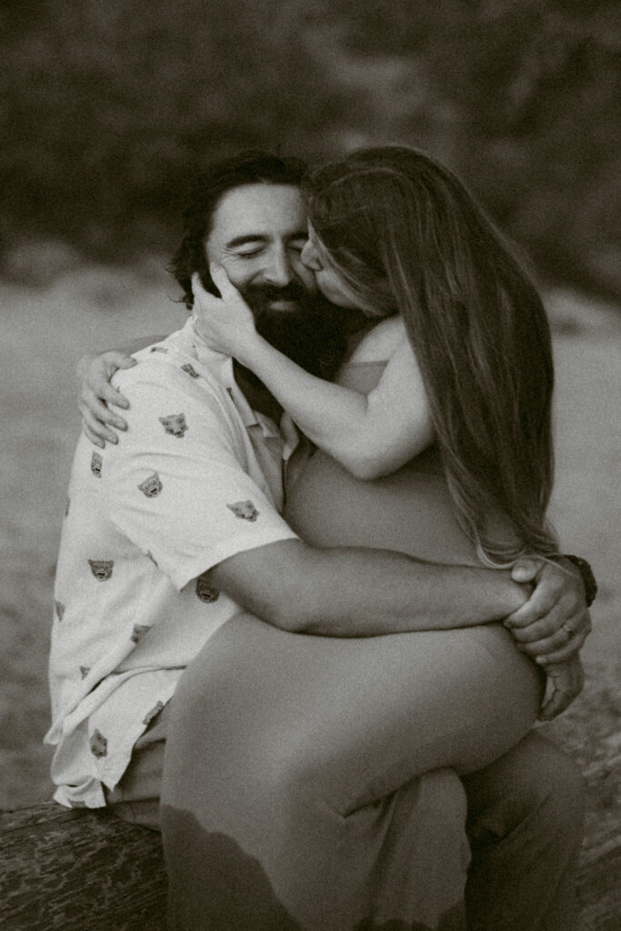 A black and white image of a young couple embracing on a log during their maternity session at Seal Rock Beach in Seal Rock, Oregon.