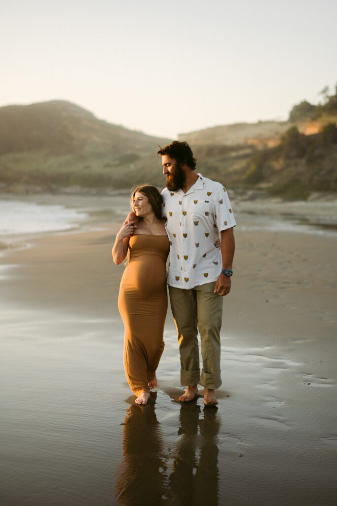 A man drapes his arm over the shoulders of his pregnant wife as they walk down the beach at Seal Rock Recreation Site for their maternity photos.