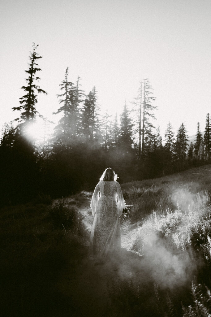 A black and white image of a bride hiking down Mount Hood on her wedding day. She's kicking up clouds of dust, staining and dress and catching in the late summer sunlight.