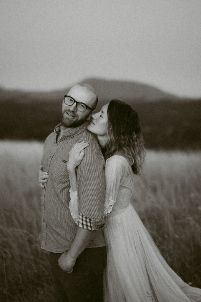 A black and white image of a woman standing on tiptoe behind a man and leaning in to kiss his cheek.