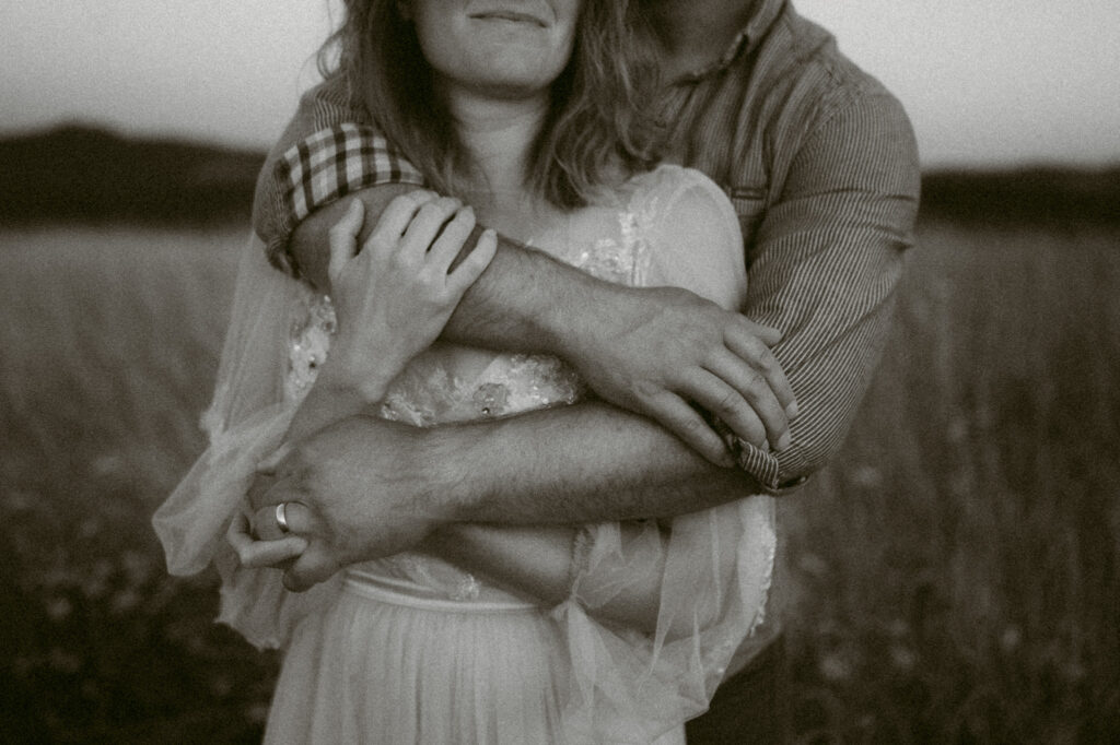 A black and white image where a man embraces a woman from behind, wrapping his arms around her chest and holding her hands.