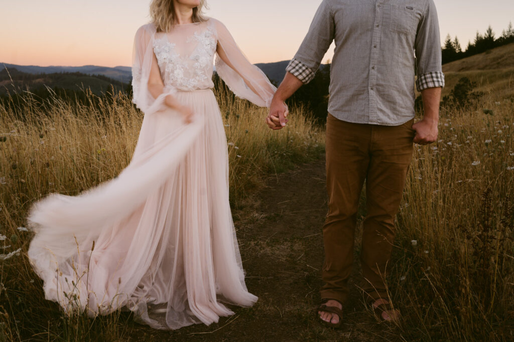 A man and woman stand slightly apart, holding hands. The woman is using her free hand to fluff the train of her gown.