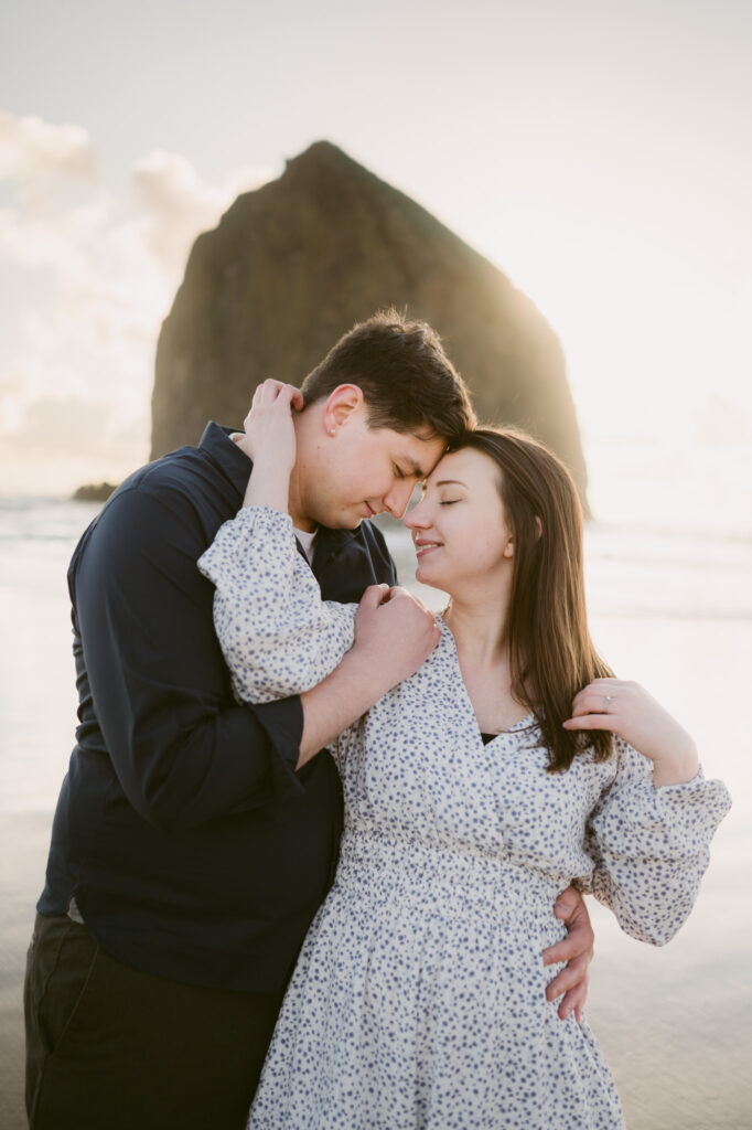 A young couple pose for their engagement photos at Cannon Beach in Oregon. The sun is setting behind Haystack Rock as they lean in, forehead to forehead.