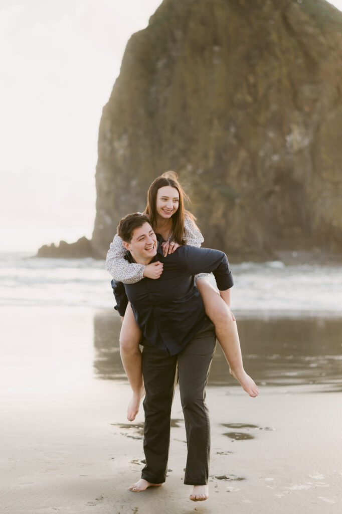 A young couple get playful during their sunset engagement session at Cannon Beach. The man is giving a piggy-back ride to the woman with Haystack Rock in the background.