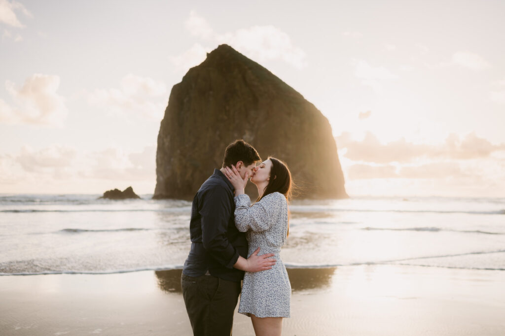 A young couple kisses in front of Haystack Rock at Cannon Beach during their sunset engagement session.