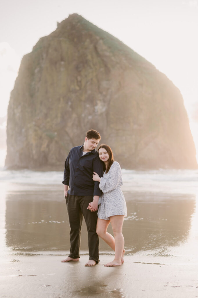 A young woman holds onto a young man's arm during their summer engagement session at Cannon Beach.