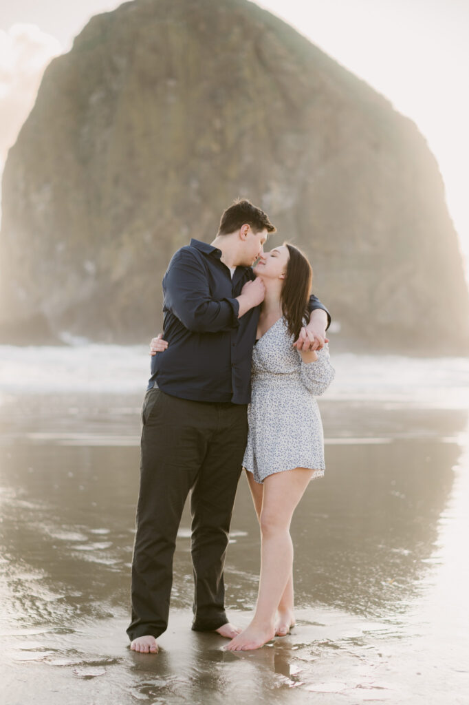 A young couple stops to kiss on Cannon Beach during their engagement session. Haystack Rock stands visible in the background.