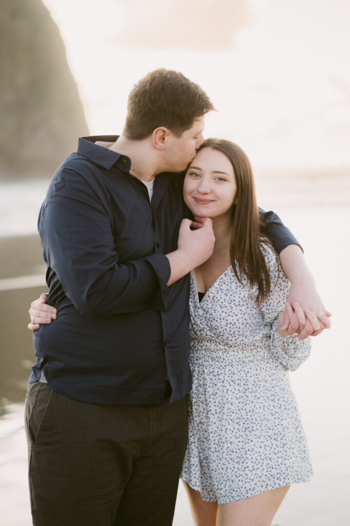 A young couple stops to kiss on Cannon Beach during their engagement session. Haystack Rock stands visible in the background. The young man has one arm draped over the woman's shoulders while the other gently lifts her chin as he kisses her temple.