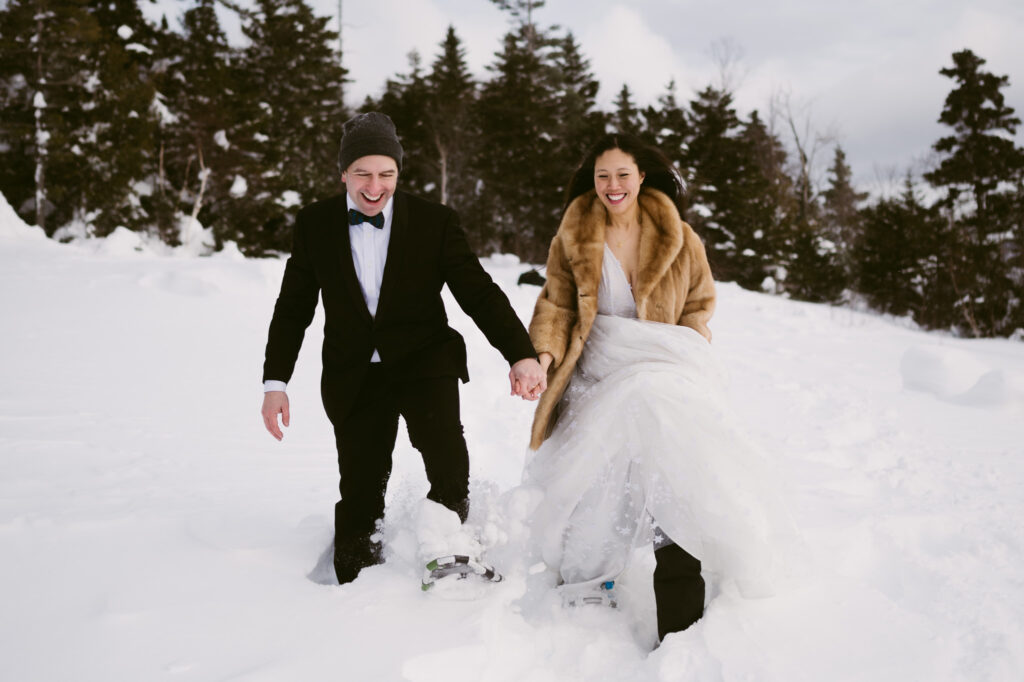 A bride and groom, bundled up and wearing snowshoes, wade through the deep snow during their winter elopement in Lake Placid, New York.