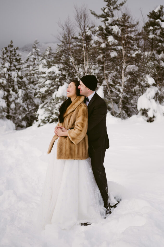 A groom holds his bride as they laugh together during their winter hiking elopement at Cobble Lookout in Wilmington, NY.