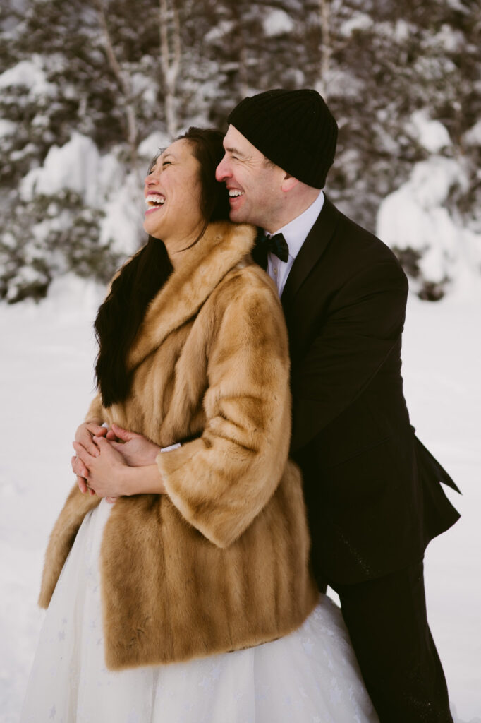 A groom holds his bride as they laugh together during their winter hiking elopement at Cobble Lookout in Wilmington, NY.