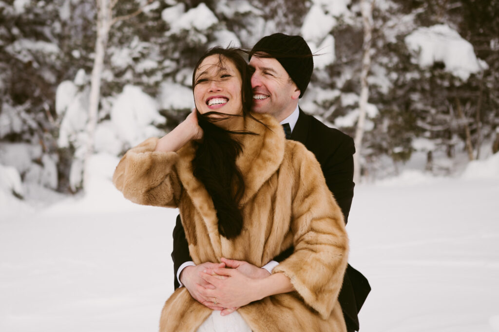 A groom holds his bride as they laugh together during their winter hiking elopement at Cobble Lookout in Wilmington, NY.