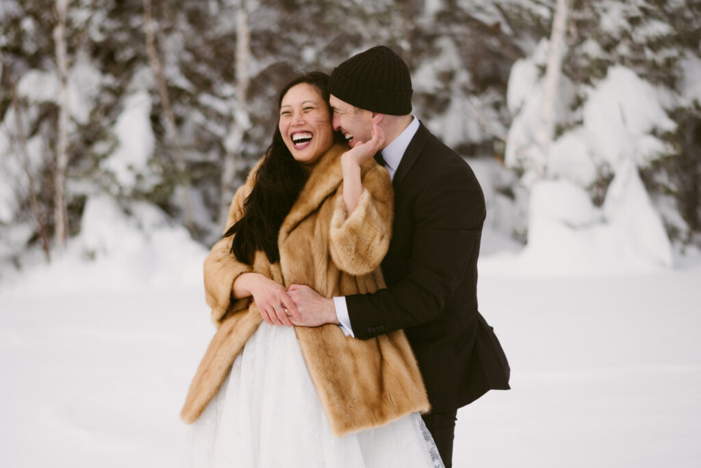 A bride leans against her groom, smiling while touching his cheek during their snowy winter wedding in upstate New York.