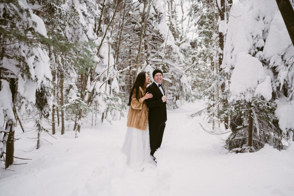 A bride wearing a fur coat hugs her groom while they pose for wedding portraits on a snowy trail.