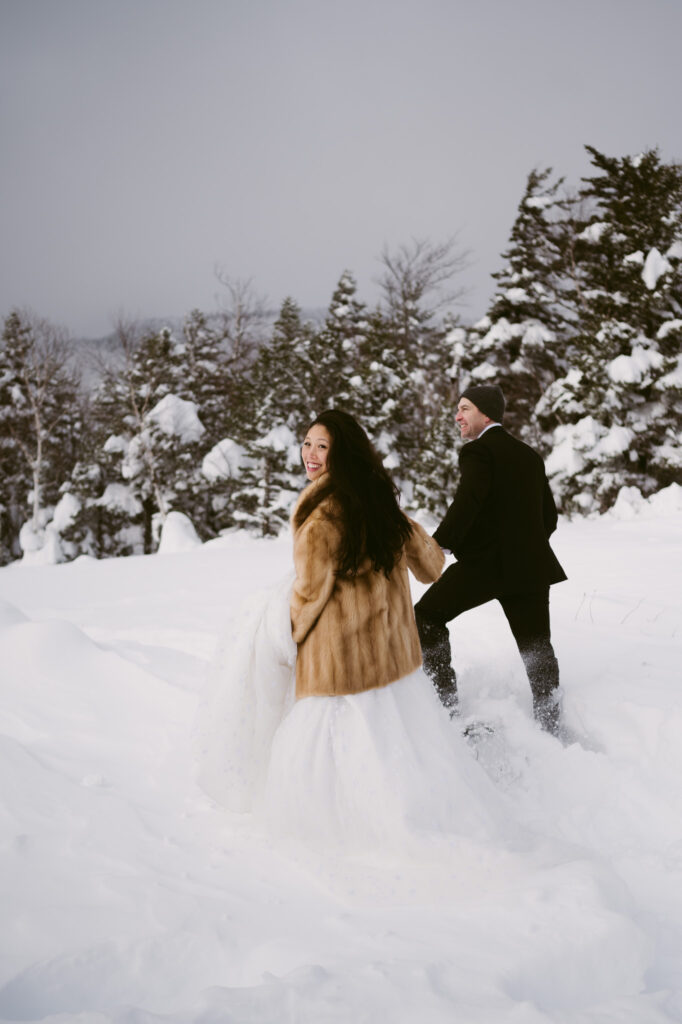 A bride and groom, bundled up and wearing snowshoes, wade through the deep snow during their winter elopement in Lake Placid, New York.