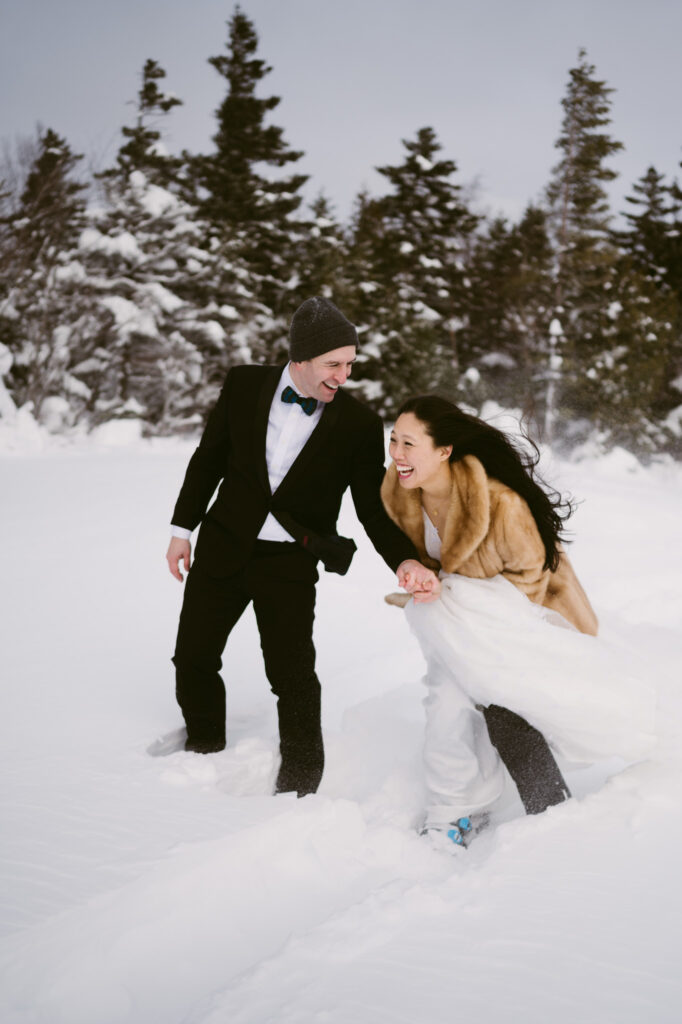 A bride and groom, bundled up and wearing snowshoes, wade through the deep snow during their winter elopement in Lake Placid, New York.