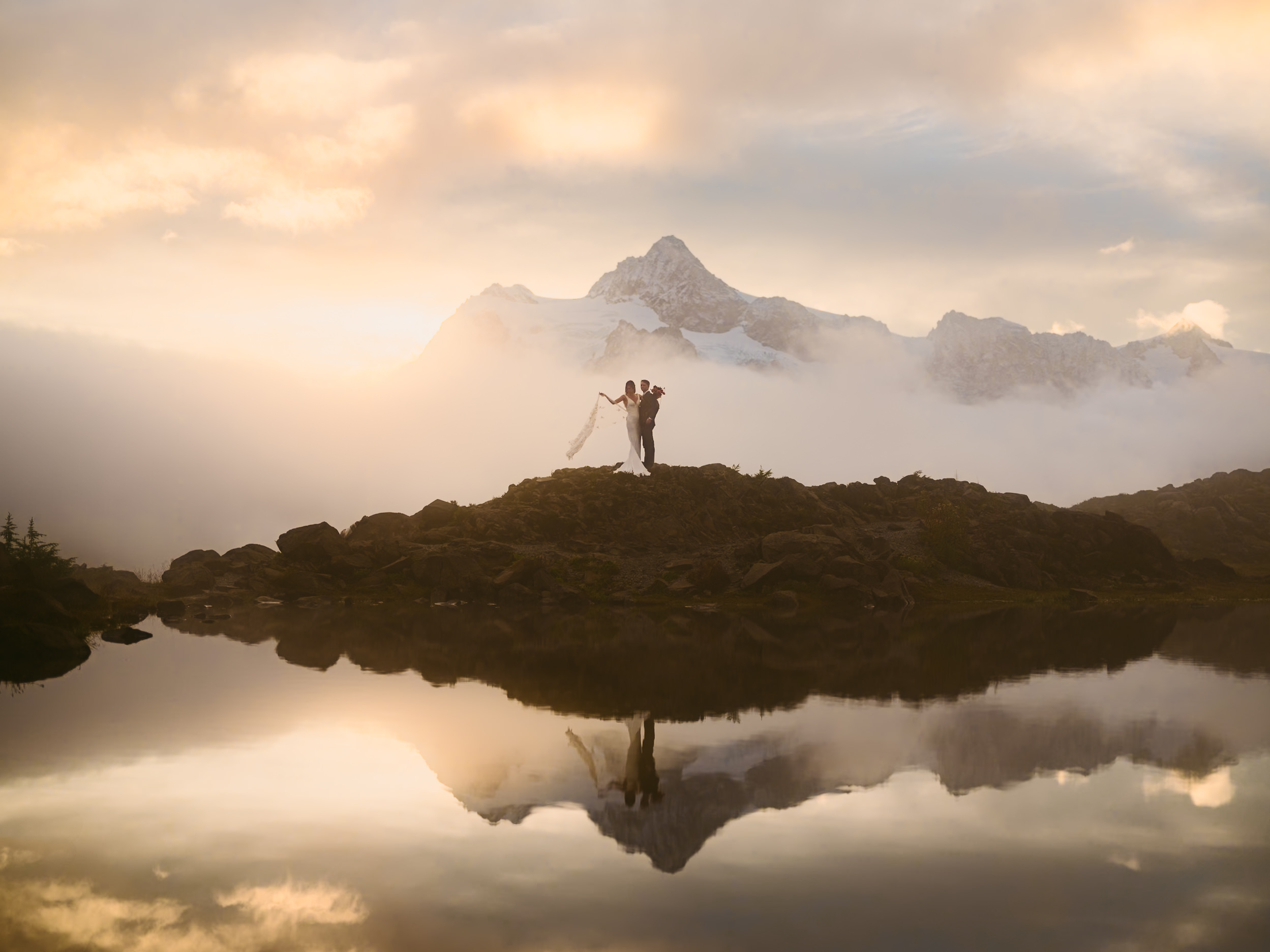 A bride and groom stand in front of Mount Shuksan at sunrise.